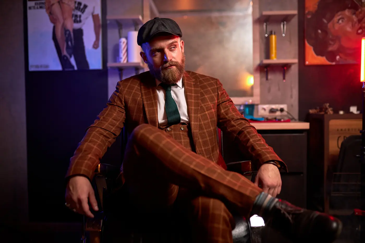 A bearded man in a checked three-piece suit and flat cap, seated in a dimly lit room