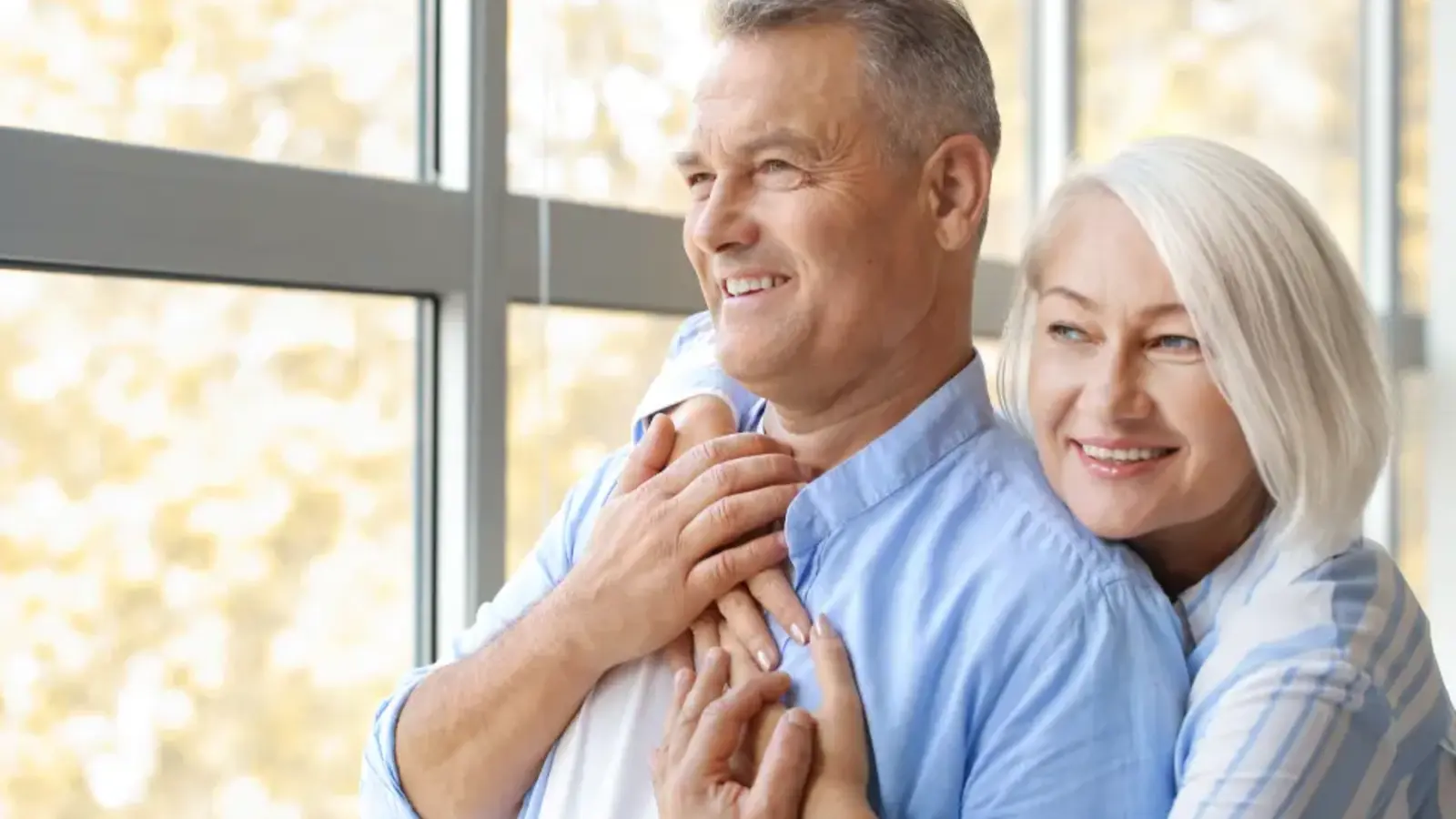 An older couple embracing and smiling by a window