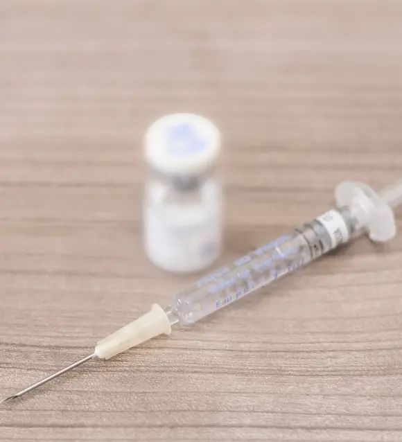 A prefilled syringe with a fine needle lying on a wooden table, a small medical vial behind it