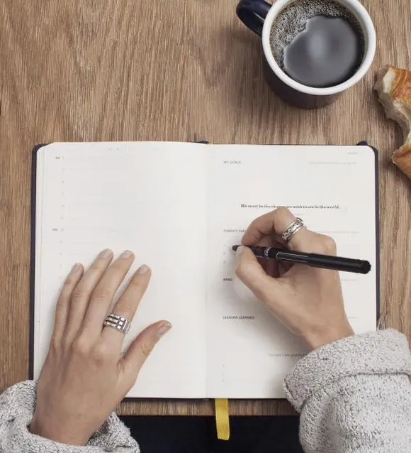 Hands writing in a notebook beside a cup of coffee